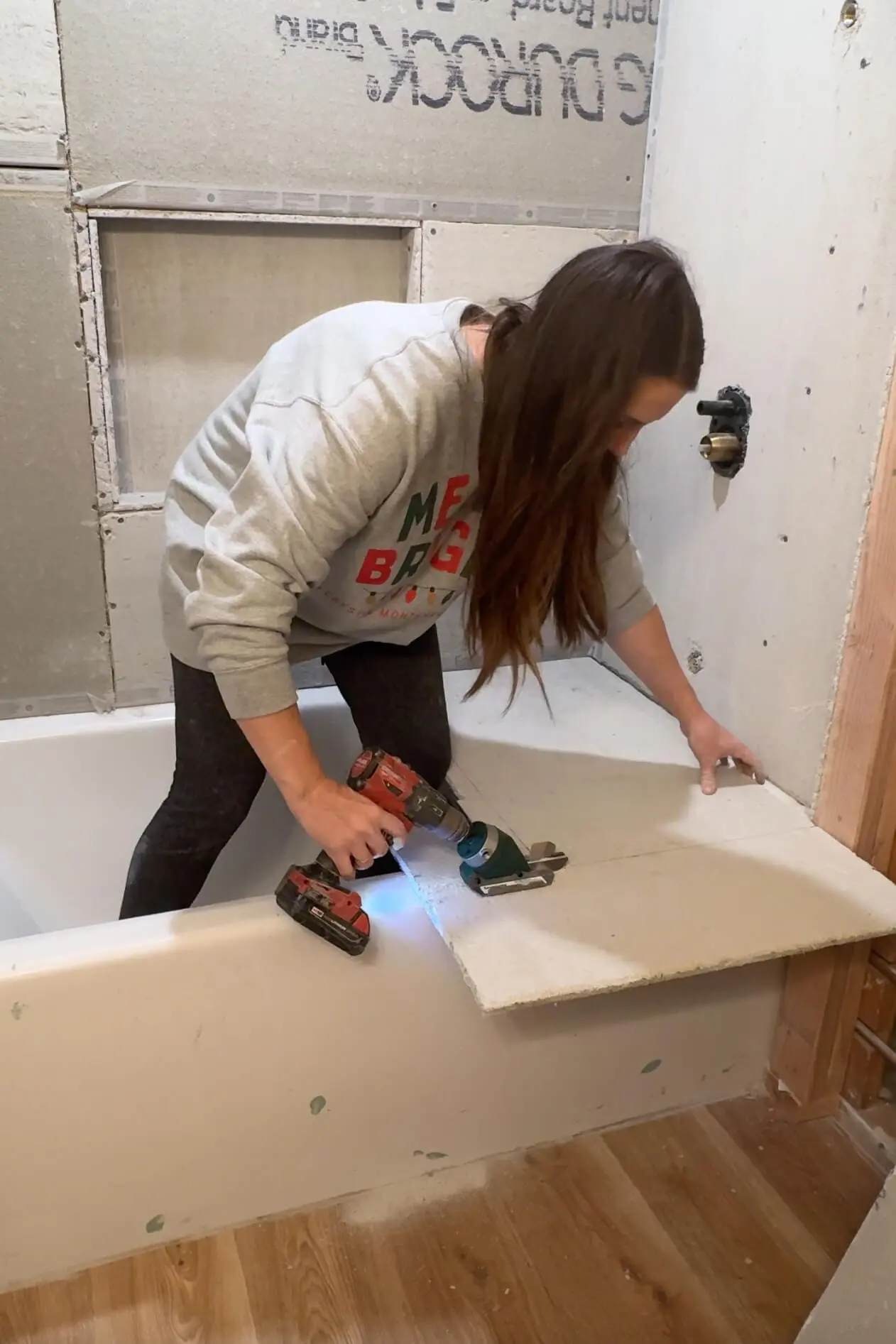 A woman wearing a gray sweatshirt is using a red power drill to cut a circular hole in a cement board resting on the edge of a bathtub during a bathroom renovation. The wall behind her has exposed cement boards and plumbing.